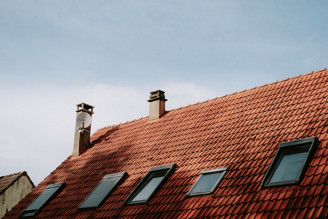 A picturesque red tiled roof featuring chimneys and skylight windows against a blue sky.